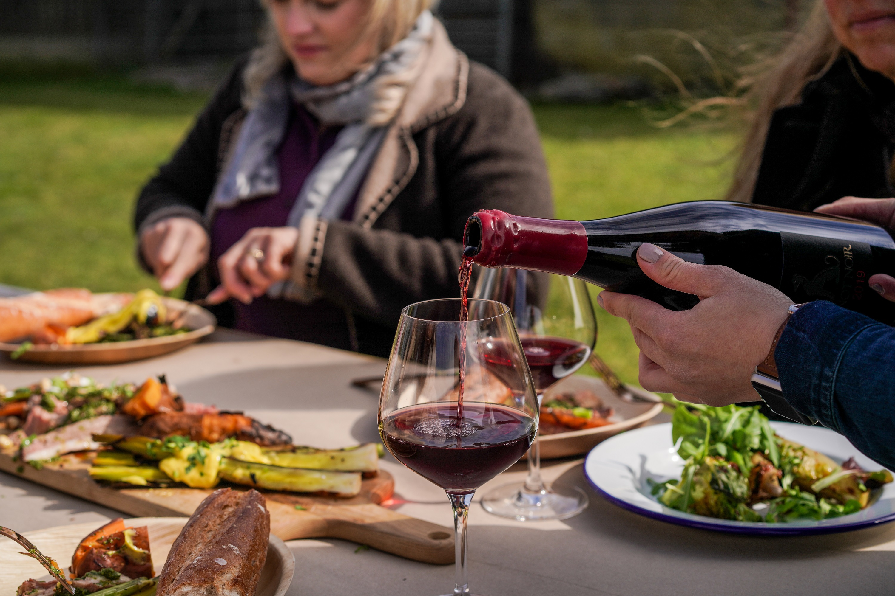 white hand pouring red wine and outdoor wooden table laden with food